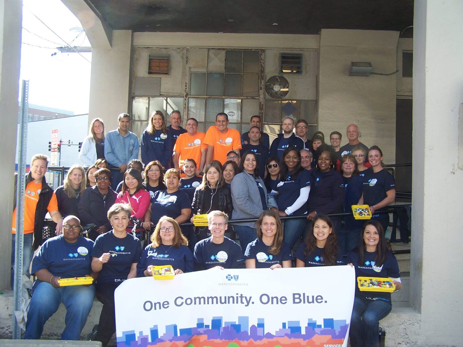 A large group of 34 corporate volunteers are smiling at the camera. They are wearing blue colored shirts which show the logo of Blue Cross Blue Shield of Massachusetts. 5 volunteers in the front are holding a large sign which states: “One Community. One Blue,” and shows the blue cross blue shield logo with a silhouette of the city.