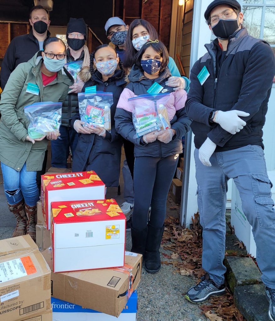 7 PMD volunteers are posing in front of the camera, displaying their project for the day of assembling snacks into packages for the street outreach program. They hold up ziplock bags that they have filled with snacks, and are wearing face masks, gloves, and PMD badges.