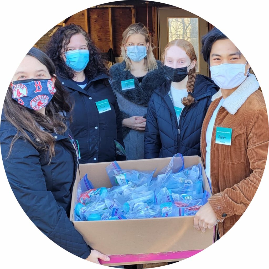 Five PMD volunteers are facing your direction, wearing face masks, PMD volunteer badges. They jointly carry a full box of packed ziploc bags that they finished assembling for the street outreach program with Pine Street Inn. Each individual bag includes items such as hand sanitizer, lip balm, lotion, facial tissue packs, and other toiletries.