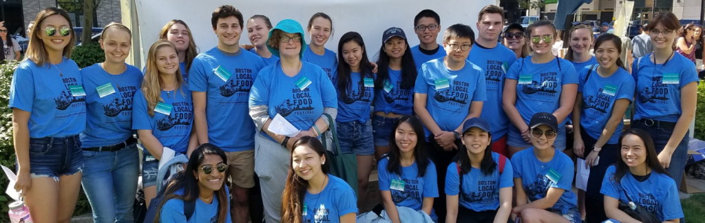 24 PMD volunteers are smiling at the camera. They are wearing PMD badges, and blue shirts which state “The Boston Local Food Festival.” The project was to help promote zero-waste, sustainable efforts.