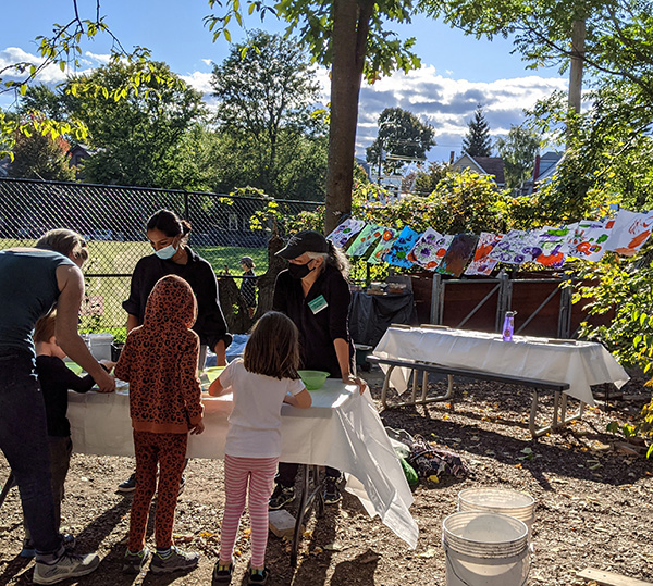 Two, masked PMD volunteers help a child and parent make prints using vegetables and vegetation. Strung behind them in the background are more than a dozen completed prints, drying clipped to a line hung between two trees at the 2021 Harvest Fest